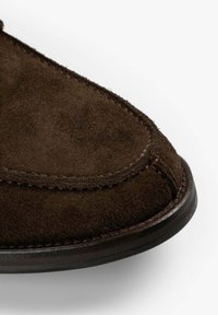 La punta del zapato de ante marrón presenta una forma redondeada, un detalle de costura a lo largo del borde y una superficie texturizada. La suela es oscura y plana.