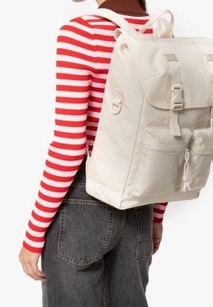 Persona que lleva una camisa de manga larga a rayas rojas y blancas y vaqueros oscuros, con una mochila grande de color blanco roto con bolsillos delanteros y cierre de hebilla.