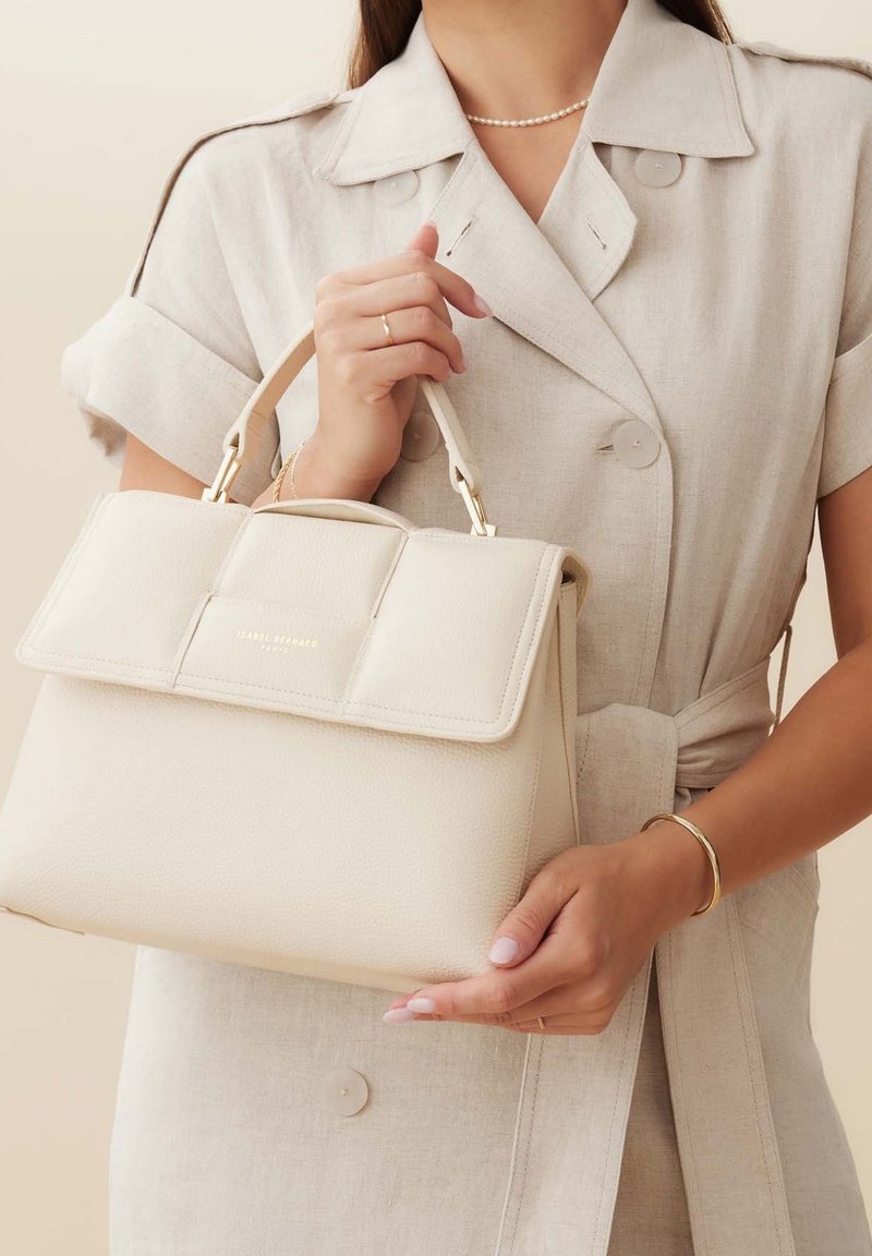 Sac à main en cuir crème avec une forme structurée, accents en métal doré et un rabat, tenu par une personne en robe boutonnée beige clair.