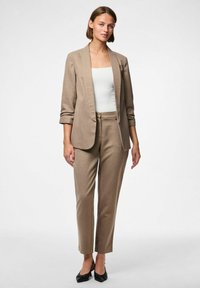 Blazer beige su misura e pantaloni coordinati realizzati in tessuto testurizzato. Il blazer presenta un colletto a scialle e una vestibilità slim; abbinato a un top bianco aderente.