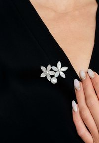 Silver floral brooch featuring clear gemstones, a pearl accent, and a textured finish, pinned on a black fabric surface.