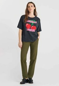 Zwart gespikkeld T-shirt met een roze en rode kersgrafiek en de tekst "Exercises Lover", gecombineerd met olijfgroene broek en zwarte schoenen.