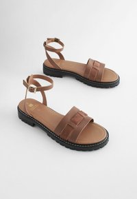 Braune flache Sandalen mit Leder- und Wildlederriemen, Knöchelschnalle und einer strukturierten schwarzen Gummisohle mit weißer Naht.