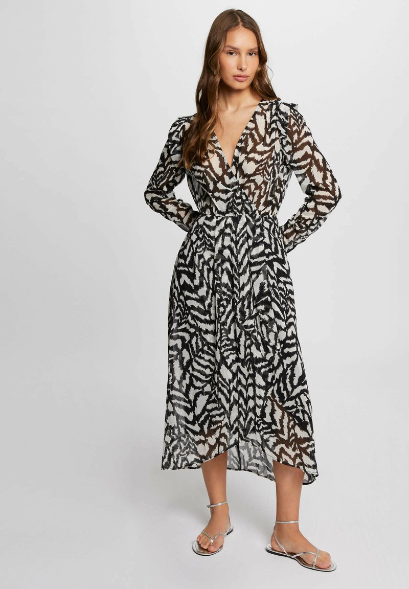 Abito lungo e trasparente con scollatura a V profonda, caratterizzato da un motivo a zigzag bianco e nero. Vita cinchata e maniche lunghe, abbinato a sandali argentati.