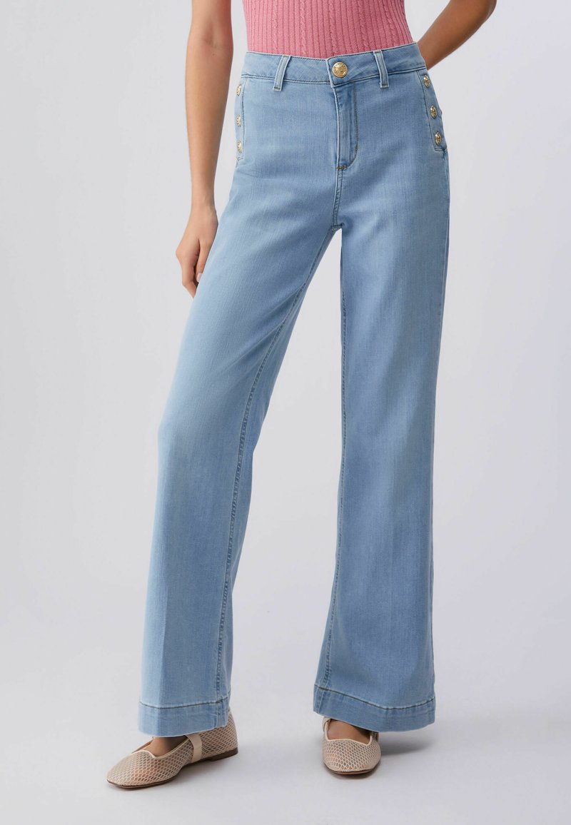 Femme portant un jean taille haute à jambes larges bleu clair avec des boutons dorés sur les hanches, assorti à un haut côtelé rose et des ballerines beiges texturées.