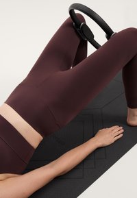 Bordo visokovratni legingi so vparjeni z črnim Pilates obročem na teksturirani črni vadbeni podlagi; roka počiva na podlagi.