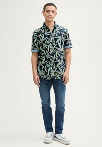 Camisa de manga corta en color navy con un patrón de hojas en verde y rosa. Con puños enrollados, combinada con jeans azules y zapatillas blancas.