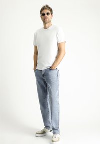 T-shirt bianco in cotone, jeans in denim blu chiaro e sneakers grigie. Gli accessori includono occhiali da sole neri e rotondi. Posizione eretta.