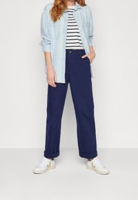 Camicia di denim blu chiaro con bottoni sopra una maglietta a righe in bianco e nero, abbinata a pantaloni larghi blu navy e sneaker bianche con dettagli marroni.