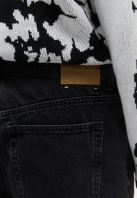 Jeans in denim nero con tasca posteriore e etichetta in pelle marrone, abbinati a un maglione a motivo bianco e nero infilato dentro.