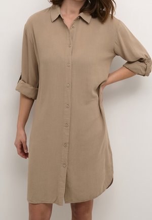 Beiges Hemdblusenkleid mit gekrempelten Ärmeln, getragen von einer Person, die vor einem einfarbigen Hintergrund steht.