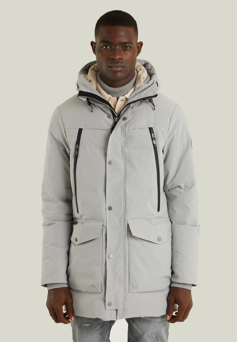 Ljust grå isolerad parkas med huva, med svarta dragkedjor, flera fickor och en slät yta. Stängs med knappar och tryckknappar.