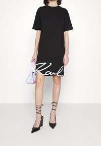 Svart kortärmad klänning med vit logotyp vid fållen, kombinerad med en handväska i pastellgradering och svarta högklackade pumps med spetsiga tår och ankelsnörning.