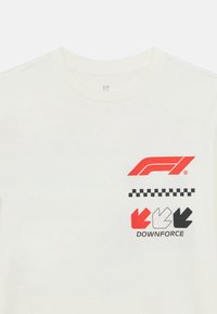 Λευκή μπλούζα με λαιμόκοψη, που φέρει κόκκινα και μαύρα γραφικά: το λογότυπο F1, σχέδιο με καρό μοτίβο και το κείμενο «DOWNFORCE» με πολύχρωμα βέλη.