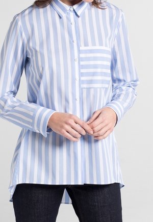 Femme portant une chemise à rayures verticales bleu clair et blanches avec une poche poitrine à rayures horizontales contrastantes.