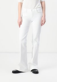 Jean flare blanc en denim, avec une fermeture à bouton à l'avant et deux poches avant, associés à des mocassins noirs.