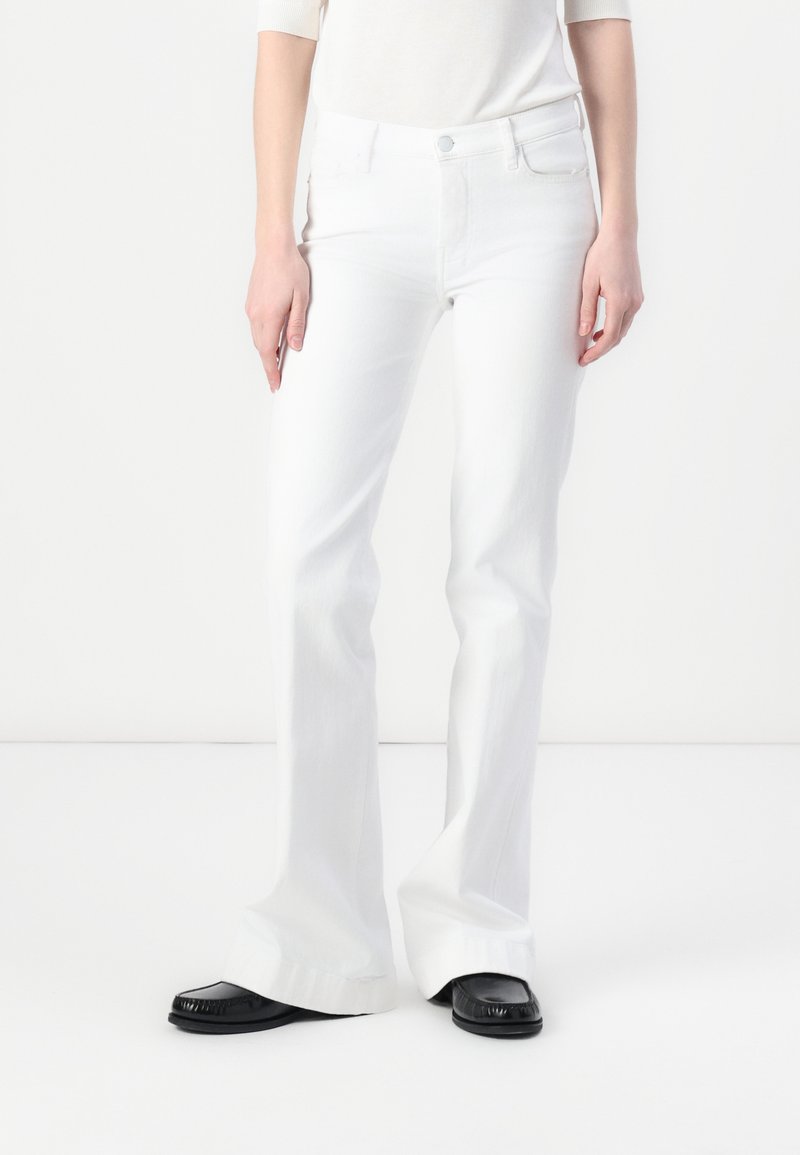 Jean flare blanc en denim, avec une fermeture à bouton à l'avant et deux poches avant, associés à des mocassins noirs.