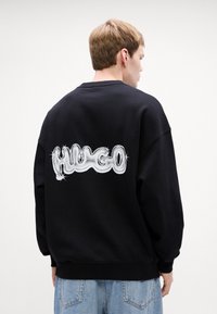 Felpa nera con un grande logo bianco lucido "HUGO" sul retro. Tessuto morbido con vestibilità rilassata, polsini e orlo a coste.