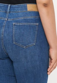 Blugi din denim albastru, cu un buzunar din spate curbat și o etichetă de marcă din piele texturată de culoare bej la talie. Design clasic cu 5 buzunare.