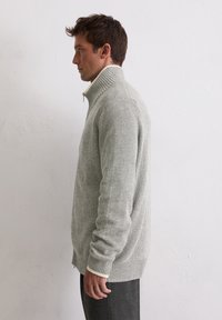Grau zip-up Pullover mit geripptem Muster, hohem Kragen und cremefarbenen Akzenten am Halsausschnitt. Seitenansicht, die das enganliegende Design zeigt.