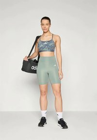 Hikiöitä harmaa urheiluliivi, jossa on kuviota, yhdistettynä vaaleanvihreisiin korkeavyötäröisiin shortseihin. Musta Adidas-käspussi ja mustat lenkkarit jalassa.