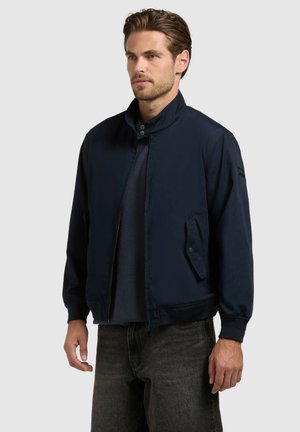 Férfi sötétkék cipzáros dzsekiben, kapcsos gallérral és zsebfedőkkel, fekete farmerral egy egyszínű háttér előtt.