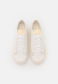 Vita canvas sneakers med gula accenter, rund tå, platta snören och varumärke på tungan och sidorna. Texturerad yta med detaljer.