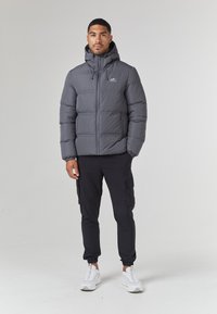 Grå pufferjakke med hette, med en frontglidelås og elastiske mansjetter; brukt med svarte cargo-bukser og hvite joggesko.