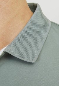 Šedé polo tričko s texturovaným límcem. Límec má jemný vzor a látka vypadá hladce a prodyšně.