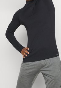 Person wearing a black long-sleeve athletic top and gray sweatpants, stretching with one arm raised and other hand on hip.