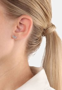 Zbliżenie na ucho blondynki z małym srebrnym kolczykiem w kształcie liścia, jej włosy związane w niskiego kucyka.