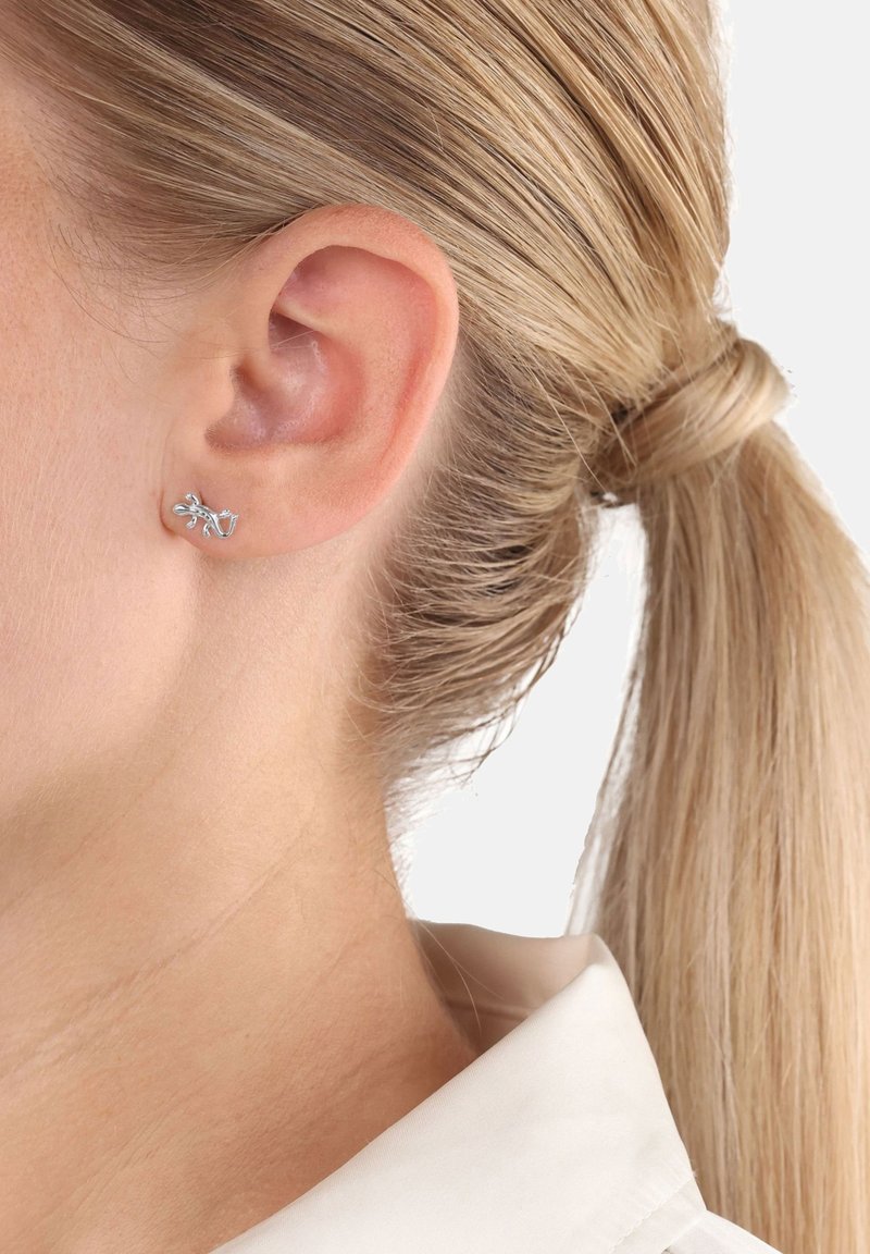 Zbliżenie na ucho blondynki z małym srebrnym kolczykiem w kształcie liścia, jej włosy związane w niskiego kucyka.