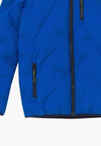 Blauwe pufferjack met zwarte ritsen aan de voorkant en zijkanten, felgroene ritstrekkers en een elastische zoom en manchetten.