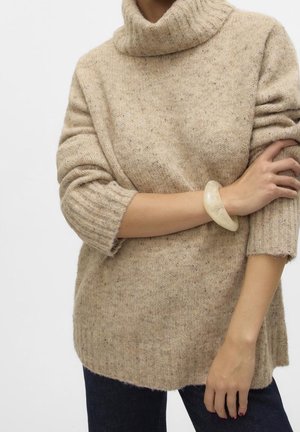 Woman wearing a beige turtleneck sweater with rolled-up sleeves and a light-colored chunky bracelet on her left wrist.