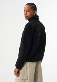 Schwarzer Fleece-Pullover mit hohem Kragen und strukturierter Oberfläche. Verfügt über eine lockere Passform und Seitentaschen, kombiniert mit hellbeigen Cargo-Hosen.