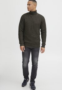 Dunkelgrüner, gestrickter Rollkragenpullover mit strukturiertem Muster, kombiniert mit grauen Slim-Fit-Jeans und dunklen Sportschuhen.