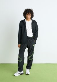 Mlad muškarac s kovrčavom kosom nosi crne Adidas trenirke s neonskim detaljima, crnu jaknu, bijelu majicu i bijele tenisice, stoji na zelenom podu.