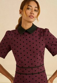 Vínová šaty s čiernymi bodkami, krátkymi puff rukávmi a čiernym golierom. Majú priliehavý pás s čiernym detailom a hladkú textúru.