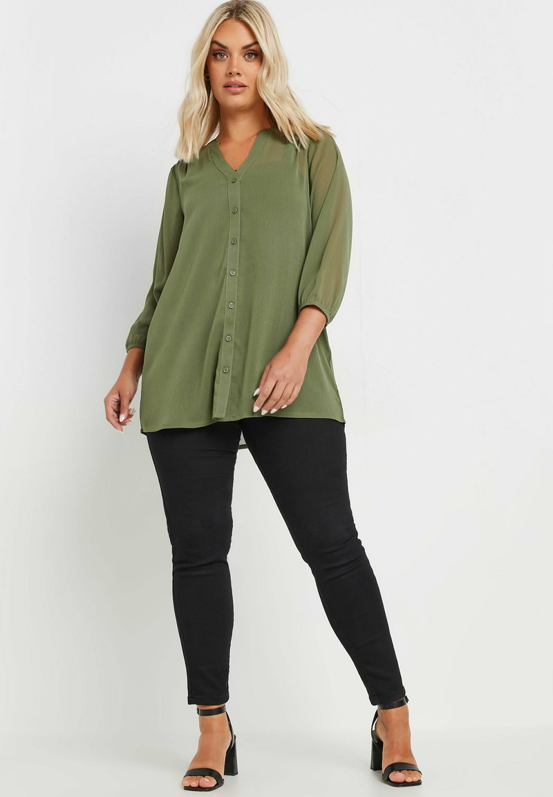 Blusa verde con bottoni, scollatura a V, maniche trasparenti e vestibilità ampia, abbinata a jeans skinny neri e tacchi neri con cinturini.