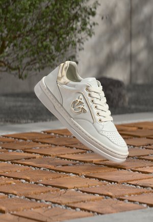 Zapatilla blanca con acentos dorados y diseño de cordones, flotando sobre baldosas de madera al aire libre, cerca de vegetación y una pared de concreto.