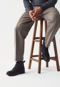 Ghete negre din piele cu design cu șireturi, având guler texturat și talpă robustă. Asortate cu pantaloni bej deschis, pe un scaun de lemn.