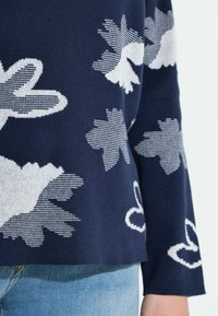 Granatowy sweter z białymi kwiatowymi i abstrakcyjnymi wzorami, dzianą fakturą, długimi rękawami oraz lekko skróconym dołem.