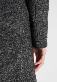 Cappotto grigio in lana con finitura testurizzata, dettagli con bottoni sui polsini e cuciture sartoriali lungo le maniche e le cuciture laterali.