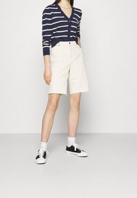 Granatowy kardigan w paski z guzikami, kremowe szorty z wysokim stanem z postrzępionymi brzegami oraz czarne sneakersy w połączeniu z białymi skarpetkami.