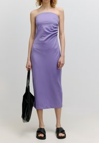 Strapless lavendelkleurige midi-jurk met gedrapeerde zijdetails. Het model houdt een zwarte tote bag vast en draagt zwarte slipper-sandalen. Gladde stofstructuur.