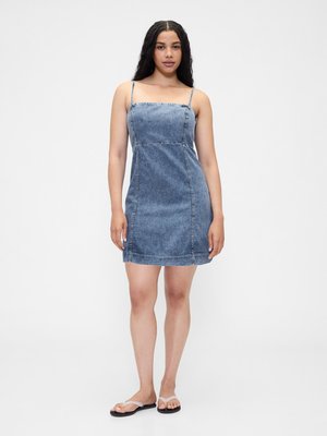 Woman standing wearing a blue denim mini dress with thin straps and flat white flip-flops against a plain white background.