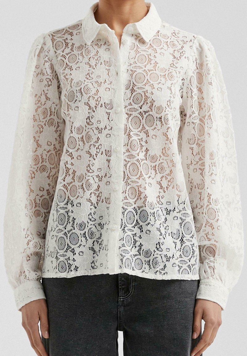 Blouse blanche en dentelle à manches longues avec motifs floraux, col, ouverture boutonnée devant et manches légèrement bouffantes, portée avec un pantalon foncé.