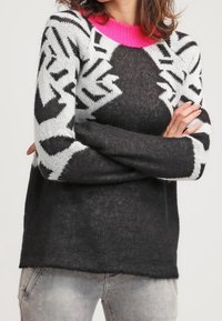 Nainen, jolla on yllään musta-valkoinen kuvioitu neulepaita kirkkaan vaaleanpunaisella kauluksella, kädet ristissä, yhdistettynä vaaleanharmaisiin haalistuneisiin farkkuihin.