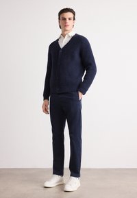 Marineblauwe gebreide cardigan met v-hals, knopen aan de voorkant, ribbelstructuur; gecombineerd met marineblauwe broek en witte sneakers.