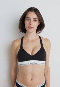 Čierny športový podprsenka s hlbokým V-krkom a širokým bielym pásom s logom "Calvin Klein". Materiál vyzerá hladko a priliehavo.